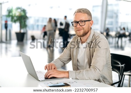 Photo of a positive attractive clever caucasian business man, with glasses, executive, programmer, company seo, manager, sits at a desk with a laptop in coworking center, looks at camera, smiling