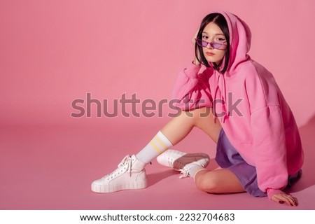 Fashionable confident woman wearing trendy pink hoodie, purple  sunglasses, mini skirt, white high top sneakers, striped socks, sitting, posing on pink background. Copy, empty space for text Royalty-Free Stock Photo #2232704683