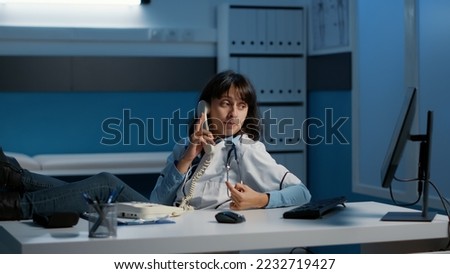 Relaxed doctor sitting with feet on desk table while talking at phone whith remote friend during night shift in hospital office. Physician woman working at medical report analyzing expertise.