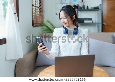 Portrait of a beautiful Asian teenage girl using her phone and computer for video conferencing sitting on the sofa at home.