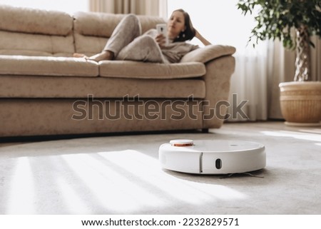 Woman is sitting on couch and operating white robot vacuum cleaner via wi-fi. Girl wearing home clothes resting relaxing lying on sofa with smartphone. Smart home