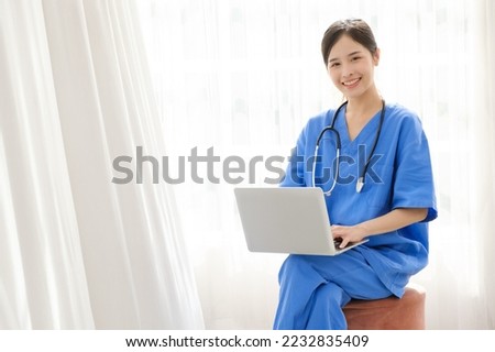 Portrait of a young Asian happy nurse wearing medical scrubs looking at the camera with a stethoscope and using a laptop while sitting with a white curtain in the background. Image with copy space.