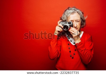 Senior woman wearing red clothes, taking pictures with an old film camera, over a red background