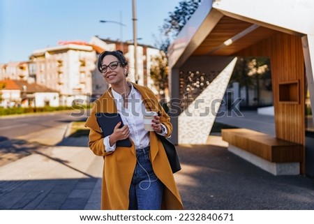 Young smiling woman waiting for a bus,uber, public transport at a stop an holding her tablet and coffee on a sunny day. Woman in jeans, coat, glasses waiting for public transport