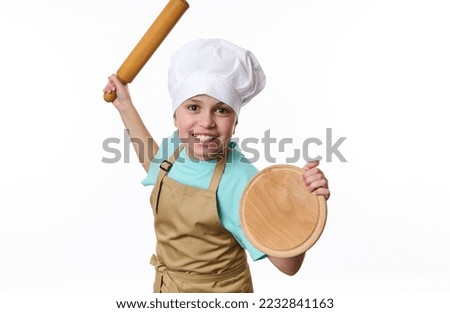 Isolated portrait on white background of a playful teenage boy, baker wearing chef's hat and apron, fighting with a wooden board and rolling pin in his hands, looking at camera. Free advertising space