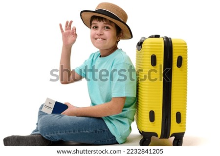 Multi-ethnic handsome teenage boy traveler wearing a turquoise t-shirt, blue jeans and straw hat, posing with yellow suitcase and boarding pass, showing OK sign looking at camera, on white background