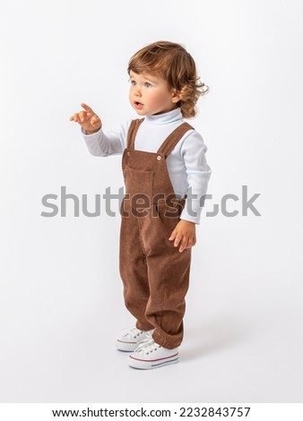 Cute Caucasian toddler 2 years old with curly hair in a brown jumpsuit, white jacket and sneakers is standing sideways and pointing his finger at something. Layout, advertising.