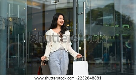Young fashionable asian woman holding shopping paper bags in hands, walking out from shopping center with smiling face