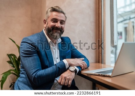 Portrait of a happy old business man sitting at his desk in the office or restaurant cafe near the window and working on a laptop. Royalty-Free Stock Photo #2232866191