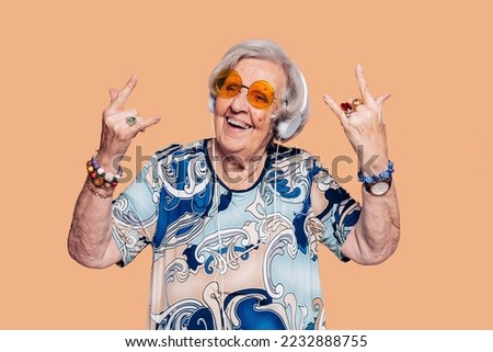 Elderly cheerful housekeeper grandmother woman 90s does horns sign gesture with hands. Stylish woman with hat, sunglasses, headphones and t-shirt over beige background.