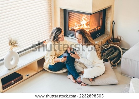 Two young women friends relaxing near fireplace, laughing, drinking wine and scrolling social media in mobile phone.