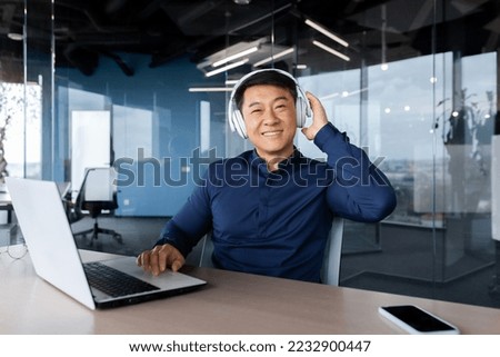 Portrait of happy asian worker in modern office, man looking at camera and smiling, boss programmer in shirt using headphones to watch video and listen to music at work.