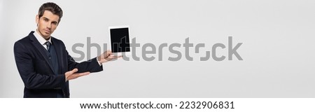 young businessman in suit holding digital tablet with blank screen isolated on grey, banner