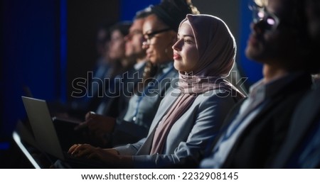 Arab Female Sitting in a Dark Crowded Auditorium at a Human Rights Conference. Young Muslim Woman Using Laptop Computer. Activist in Hijab Listening to Inspiring Speech About Global Initiative. Royalty-Free Stock Photo #2232908145