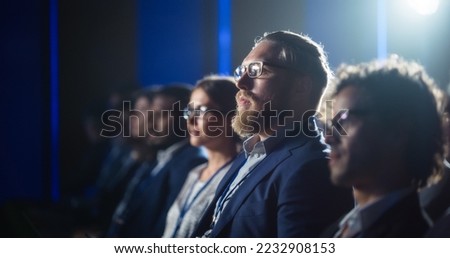 Young Bearded Man Attending a Political Conference, Sitting in Crowded Hall. Journalist Taking Notes and Writing an Article on Laptop Computer. Writer Listening to a Motivational Speaker. Royalty-Free Stock Photo #2232908153