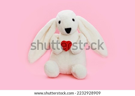 children's toy a white hare with long ears holds a red heart in its paws, isolate on a pink background