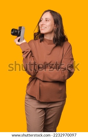 Vertical shot of a young woman holding a vintage camera and looking aside over yellow background.