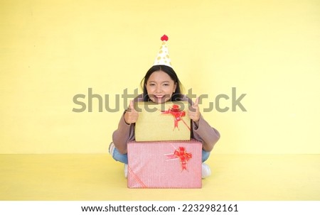 Happy young girl wear party hat and hold gift box on yellow background.