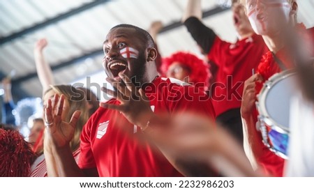 Sport Stadium Big Event: Handsome Expressive Black Man with Painted face Jumping Up and Cheering Goal Score. Crowd of Fans Shout for Red Soccer Team to Win. People Celebrate Championship Victory Royalty-Free Stock Photo #2232986203