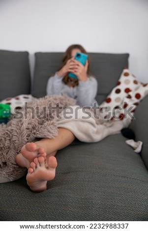 Young girl, teen, preteen, sitting on a large long canape sofa. Visibly bored, center of focus is on her feet that are in front of the camera. Spending time sick, home, being isolated, sad, lazy. Mess