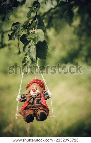 A cheerful stylish clown toy in a brown-red suit rides on a swing in the forest near a tree and grass for the joy and festive mood of a child. Image for your creativity, design or illustrations.