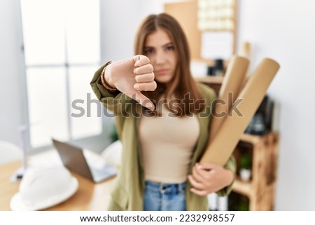Young brunette woman holding paper blueprints at the office with angry face, negative sign showing dislike with thumbs down, rejection concept 