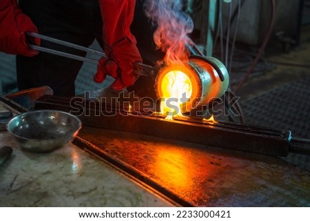 The master's hands in special red refractory mittens pour molten gold from a refractory glass into a mold. Royalty-Free Stock Photo #2233000421