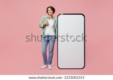 Full body young fun woman wear green shirt white t-shirt near big huge blank screen mobile cell phone smartphone with workspace mockup area show thumb up isolated on plain pastel light pink background