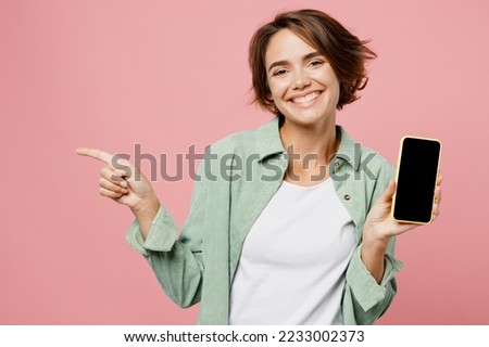 Young happy woman she wear green shirt white t-shirt hold in hand use mobile cell phone with blank screen workspace area point index finger aside isolated on plain pastel light pink background studio