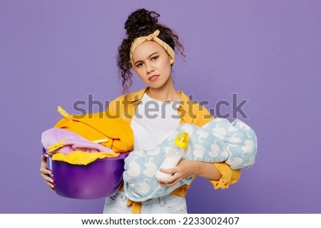 Young sad housekeeper woman wear yellow shirt tidy up hold newborn baby basin with clothes for laundry doing everyday routine isolated on plain pastel light purple background studio. Housework concept Royalty-Free Stock Photo #2233002407