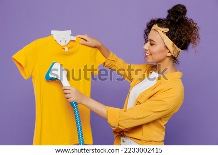 Sideways young housekeeper woman wear yellow shirt hold electrical stramer for dry-cleaning and ironing t-shirt isolated on plain pastel light purple background studio. Housework tidying up concept