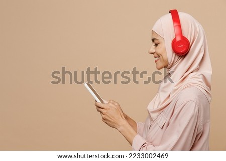 Side view young arabian asian muslim woman she wear abaya hijab pink clothes headphones listen music isolated on plain pastel light beige background. People uae middle eastern islam religious concept