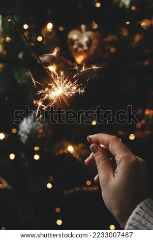 Burning sparkler in female hand on background of christmas tree lights in dark room. Happy New Year! Hand holding firework against stylish decorated tree with illumination. Atmospheric time Royalty-Free Stock Photo #2233007487