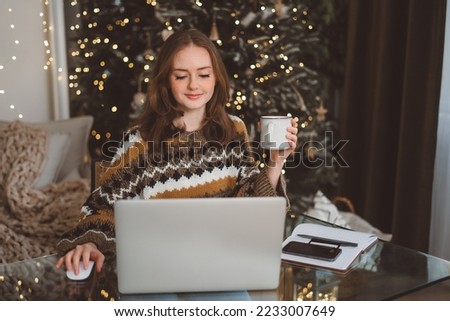 Portrait of smiling woman working on laptop or shopping online, buying new year presents with decorated Christmas tree on background, typing on keyboard. High quality photo