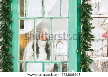 blurry portrait through window of young beautiful brunette woman in country house in winter, suburban area with stylish decor, atmosphere of Christmas and New year celebration background