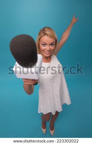 Half-length portrait of lovely smiling fair-haired TV presenter wearing pretty white dress holding a microphone wanted to interview us. Top view. Isolated on blue background