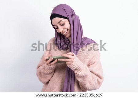 muslim woman wearing hijab and knitted sweater over white background holding in hands cell playing video games or chatting