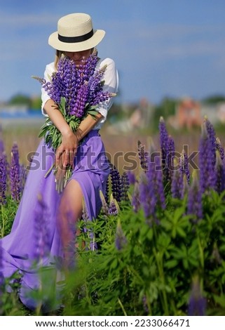                               a woman in a hat stands in the middle of a field and holds a bouquet of violet wild flowers in her hands. the girl covers her face with a bouquet looks down. Picture 