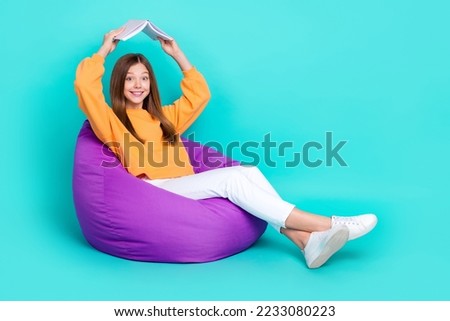 Full length photo of sweet cute schoolgirl wear orange sweatshirt bean bag showing book roof empty space isolated turquoise color background