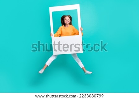Full length photo of sweet cute school girl dressed orange hoodie jumping high holding paper frame isolated teal color background