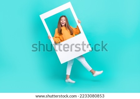 Full length photo of pretty excited schoolgirl wear orange sweatshirt holding paper photo frame isolated turquoise color background