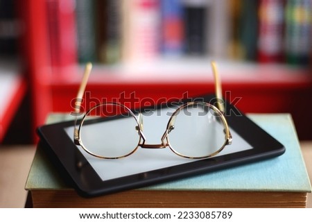 Stack of hardcover books, e-reader and reading glasses on the table. Bookshelf in the background. Selective focus.