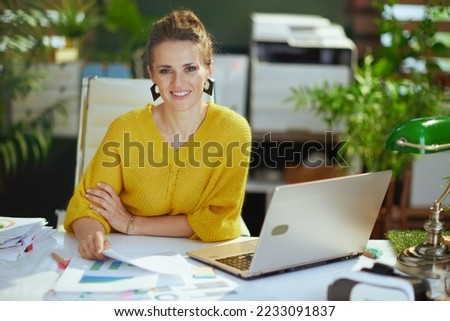 happy elegant 40 years old small business owner woman in yellow sweater with laptop working with documents in the modern green office. Royalty-Free Stock Photo #2233091837