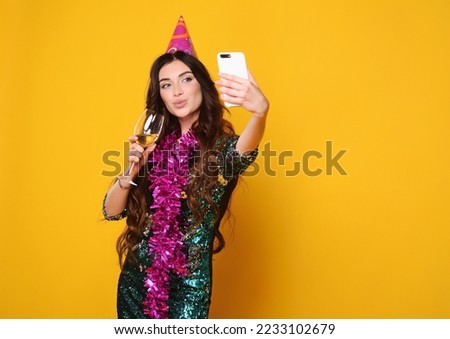 young pretty woman in a pink party hat and pink boa holds a glass of champagne in her hands, smiles and holds a smartphone in her hands