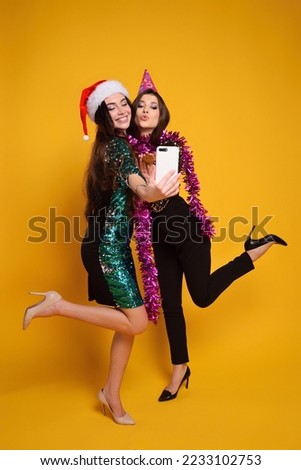 two young pretty women in sparkly cocktail dresses, party hat and santa hat and pink boa smile and take selfie on smartphone.