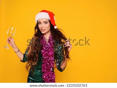 pretty woman in green sparkly cocktail dress wearing santa hat holding pink boa and champagne glass smiling on yellow background
