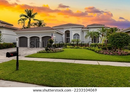 Facade of an elegant house with gray walls with white details, a red tiled roof, a front garden with abundant tropical plants, palm trees, short grass, sidewalk, driveway and garage Royalty-Free Stock Photo #2233104069