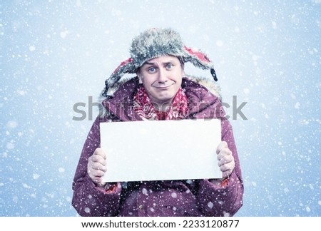 Pitiful frozen man in winter clothes holds an empty white poster in his hands, it is snowing on a blue background