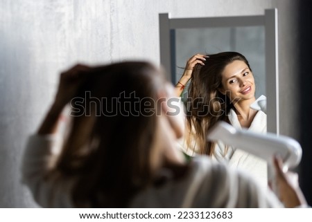 Beautiful young woman using hair dryer near mirror at home Royalty-Free Stock Photo #2233123683