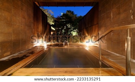 A Japanese-style open-air Kashikiri Buro (a charter hot-spring bath) in a Ryokan, with a wooden bathtub illuminated at blue dusk and a relaxing vibe in the natural setting of a green forest Royalty-Free Stock Photo #2233135291
